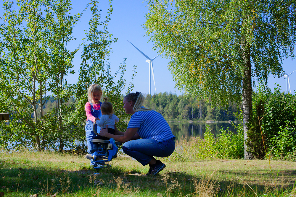 En barnfamilj ute i naturen i förgrunden och ett vitt vinkdraftverk i bakgrunden.
